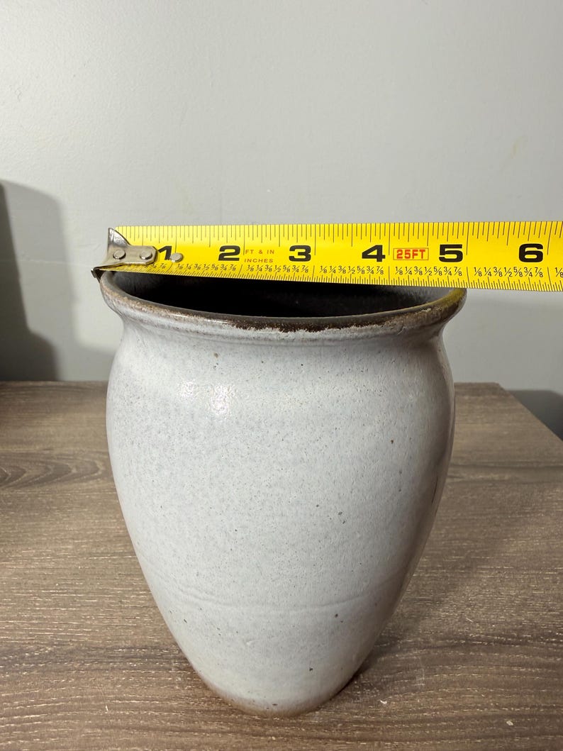 May include: A handmade, off-white ceramic vase with a dark brown rim. The vase is approximately 6 inches tall and has a speckled texture. The vase is sitting on a wooden surface.