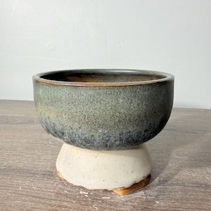 May include: A ceramic bowl featuring a speckled dark green and charcoal gray glaze on the upper portion, transitioning to a smooth, unglazed off-white base with a wide, flared pedestal foot and a visible brown clay rim at the bottom edge.
