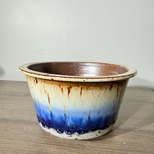 May include: A wide, round ceramic bowl featuring a multi-colored drip glaze. The interior is a deep brown, while the exterior transitions from a cream-colored upper rim down through a light blue middle section to a dark blue base with speckles.