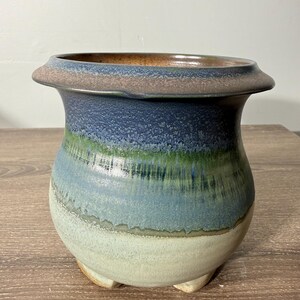 May include: A handcrafted ceramic planter with a rounded body and three small feet. The planter features a color gradient, transitioning from a light beige base to shades of blue, green, and brown at the rim. The rim has a flared design.