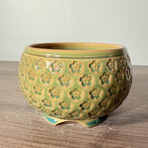 May include: A round ceramic planter with a green and yellow glaze featuring a repeating embossed flower pattern across the exterior. The bowl-shaped vessel has a smooth rim and rests on three small, integrated feet at the base.