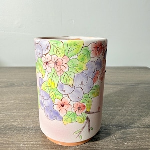 May include: A cylindrical ceramic cup featuring a hand-painted design of light purple berries, green leaves, and small pink flowers against a soft pink background. The base of the cup shows the natural terracotta color of the unglazed clay material.