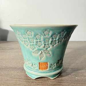 May include: A turquoise ceramic planter with a flared rim and a tapered body. The exterior features a raised floral pattern and a central leaf motif. A small, square, orange-brown stamp with embossed characters is visible near the base.