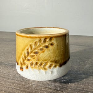 May include: A cylindrical ceramic cup featuring a two-tone glaze with a glossy golden-brown upper section and a white lower section. The exterior displays a raised, embossed wheat stalk pattern and a distinct crackle finish throughout the glaze.