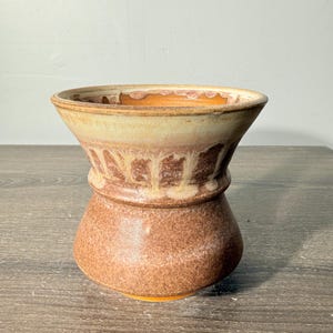 May include: A ceramic vessel with a wide, flared rim and a tapered waist. The exterior features a textured, speckled brown base glaze that transitions into a lighter cream-colored glaze with vertical drip patterns around the upper section.