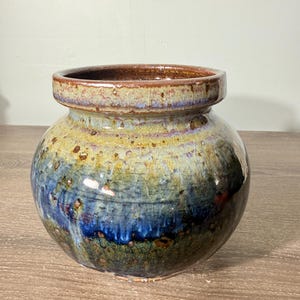 May include: A round ceramic vase with a wide, thick rim and a crackled glaze finish. The surface features a blend of earthy tones, transitioning from a golden-tan upper section to a deep, vibrant blue and dark green base with brown speckles.