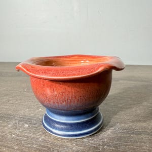 May include: A ceramic bowl with a wide, ruffled rim featuring a gradient color transition from a vibrant orange-red at the top to a deep, solid blue at the circular pedestal base. The interior of the bowl shows a warm orange hue.