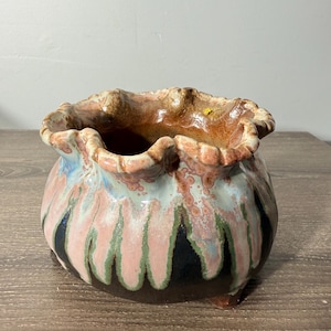 May include: A ceramic planter with a ruffled rim. The planter features a color palette of brown, pink, black, and green. The base has three small feet. The planter is suitable for indoor or outdoor use.