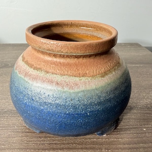 May include: A round ceramic pot with a wide opening and a textured surface. The pot features a color gradient, transitioning from a deep blue base to a reddish-brown rim, with bands of green and pink in between. The pot has three small feet.