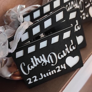 Peut inclure: Clapet noir et blanc avec le texte "Cathy & David 22 Juin 24" en lettres blanches. Le clap a une forme de cœur blanc en bas à droite.