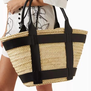 以下が含まれることがあります： 黒い革のストラップが付いた、織り込まれた麦わらトートバッグ。 バッグは長方形で、黒いハンドルが2つ付いています。 バッグは、黒い花の刺繍が施された白いドレスを着た人が持っています。