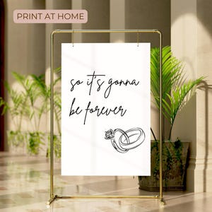 May include: A white printable sign with the text "so it's gonna be forever" and a black line drawing of two intertwined rings. The sign is on a gold stand.