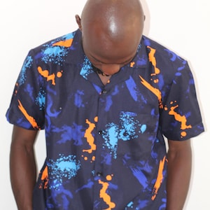 Puede incluir: Una camisa de manga corta con botones, de fondo azul y con patrones abstractos de color naranja y azul.