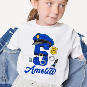 May include: White t-shirt with a blue number 5, a police hat, handcuffs, a flashlight, and a walkie-talkie. The text "Amelia" is written in blue below the number.