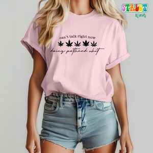 May include: A light pink t-shirt with the text "can't talk right now" above five black cannabis leaves and the phrase "doing pothead shit" in a script font.