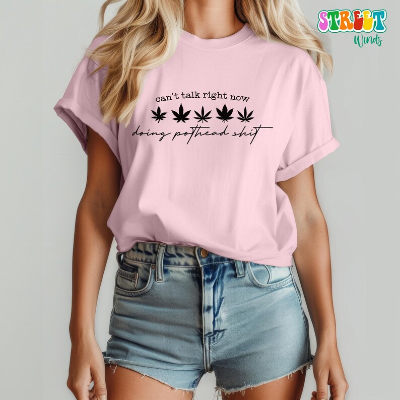 May include: A light pink t-shirt with the text "can't talk right now" above five black cannabis leaves and the phrase "doing pothead shit" in a script font.