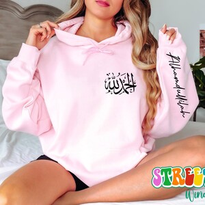 May include: Pink hoodie with a black Arabic calligraphy design on the front and the text "Alhamdulillah" on the sleeve.