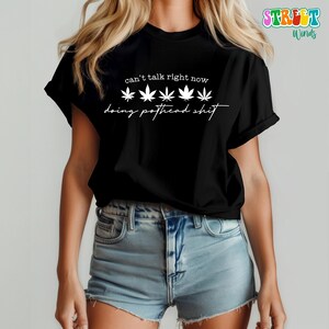 May include: Black t-shirt with white text that reads "can't talk right now doing pothead shit." The design includes five cannabis leaf illustrations. The shirt is worn with blue denim shorts.