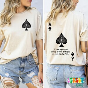May include: A beige t-shirt with a black ace of spades graphic on the front and the text "It's not about the cards you're dealt but how you play them" and a black ace of spades graphic on the back.