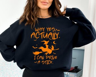 May include: Black sweatshirt with orange text that reads "WHY YES, ACTUALLY. I CAN DRIVE A STICK." The graphic features a witch on a broom with a cat and bats. A Halloween-themed design.