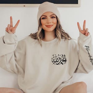 May include: A beige sweatshirt with the Arabic phrase "Allah" and the phrase "Alhamdulillah" on the sleeve. The model is wearing a beige beanie.