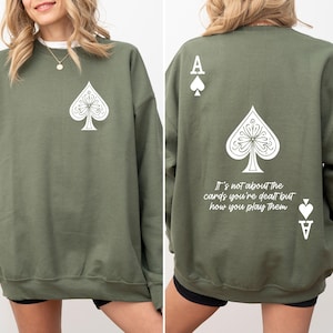May include: A green sweatshirt with a white ace of spades design on the front and back. The back design includes the text "It's not about the cards you're dealt but how you play them."