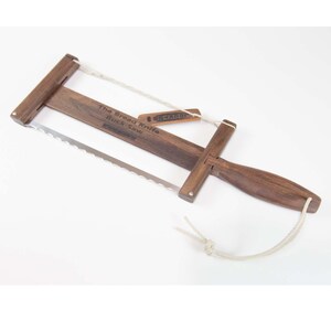May include: A wooden bread knife with a buck saw design. The knife features a dark brown wooden frame, a serrated stainless steel blade, and a white rope. The text "The Bread Knife Buck-Saw" is printed on the wood.
