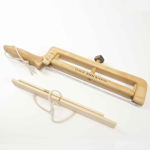 May include: A wooden cheese slicer with a handle and an adjustable thickness setting. The slicer has a knob to adjust the thickness of the slices. The text "SLICE THICKNESS" is printed on the slicer.