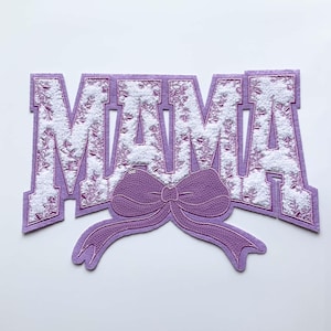 May include: A purple felt applique with the word "MAMA" in a varsity-style font. The letters are filled with white floral embroidery. A sequined purple bow is below the word. The applique is likely for clothing or crafts.
