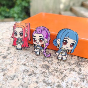 Peut inclure: Trois épingles en émail colorées représentant des personnages de style anime avec des cheveux rouges, violets et bleus. Chaque épingle a un design unique, avec des accents dorés et des expressions faciales détaillées. Les épingles sont exposées sur une surface orange.