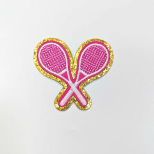 May include: An embroidered patch featuring two crossed tennis rackets in pink and white. The rackets have a textured design and are outlined in white. The patch is surrounded by a gold glitter border, creating a decorative accessory.
