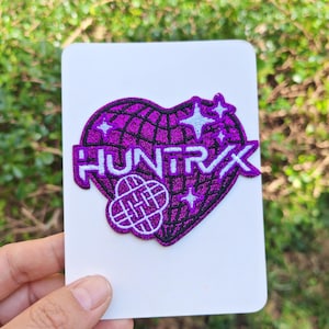 Puede incluir: Un parche bordado morado con la palabra "HUNTRX" en letras blancas, sobre un corazón con un patrón de cuadrícula. El parche incluye diseños de estrellas y globos. El parche está sobre una tarjeta blanca, sostenida por una mano.