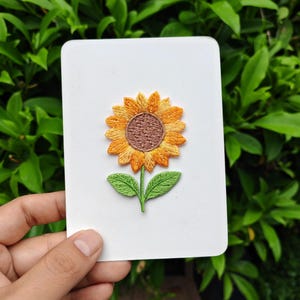 May include: A white card featuring an embroidered sunflower. The sunflower has yellow petals, a brown center, and green leaves and stem. The card is held against a backdrop of green foliage.