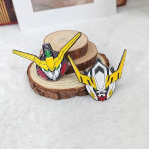 May include: Two enamel pins featuring robot head designs. One pin has a yellow, green, red, and black color scheme, while the other has a white, yellow, and red color scheme. The pins are displayed on a wooden slice.