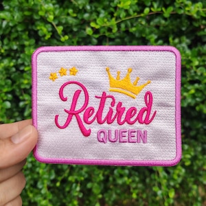 Könnte beinhalten: Rechteckiger Aufnäher mit rosa Rand und hellrosa Hintergrund. Der Aufnäher zeigt die Worte "Retired QUEEN" in rosa Schrift, eine goldene Krone und drei goldene Sterne.