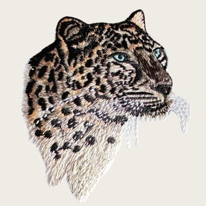 Puede incluir: Primer plano de un parche bordado con un leopardo de las nieves con ojos azules. El parche es blanco con bordados negros y marrones.