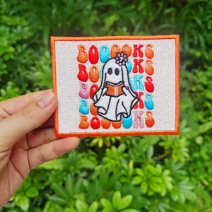 Könnte beinhalten: Ein weißer gestickter Aufnäher mit einem Geist, der ein Buch liest. Der Hintergrund ist ein sich wiederholendes Muster des Wortes "BÜCHER" in Orange, Rot, Blau und Rosa.