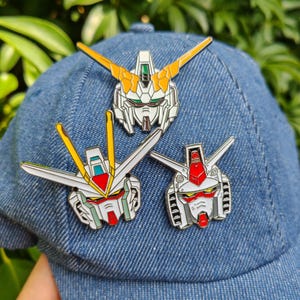 May include: Three colorful Gundam robot head pins are displayed on a denim baseball cap. The pins feature detailed designs with white, red, yellow, and green accents. The pins are attached to the cap, showcasing a fan's love for the anime series.