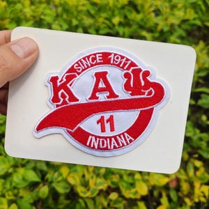 May include: A white patch featuring a red and white embroidered design. The design includes the letters "KA" in red, the number "11", and the word "INDIANA". The text "SINCE 1911" is also included in the design.