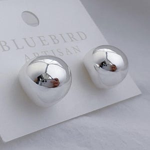 Puede incluir: Un par de pendientes de botón plateados brillantes, de forma redondeada e irregular. Los pendientes se muestran en una tarjeta blanca con el texto "BLUEBIRD ARTISAN". Los pendientes reflejan el entorno.