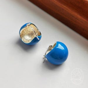 May include: A pair of blue enamel hoop earrings with a silver-tone interior and clasp. The earrings have a rounded, half-sphere shape and a glossy finish. The earrings are displayed on a white surface.