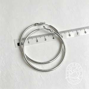 May include: A pair of silver hoop earrings. The earrings are round and have a simple, polished design. A ruler is placed next to the earrings, showing measurements in inches. The earrings are on a white surface.