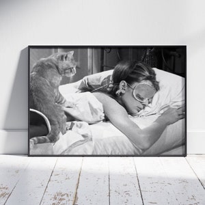 May include: A black and white photograph of a woman sleeping in bed with a cat sitting on the edge of the bed. The woman is wearing a sleep mask and has her hair pulled back. The cat is looking at the camera.