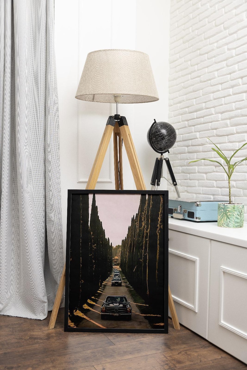 May include: A black and white framed print of a vintage photograph of a line of black cars driving down a road lined with tall trees.