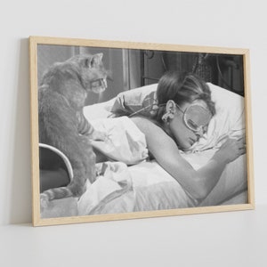 May include: A black and white photograph of a woman sleeping in bed with a cat sitting on the bed next to her. The woman is wearing a sleep mask and has her hair pulled back. The cat is looking at the camera.