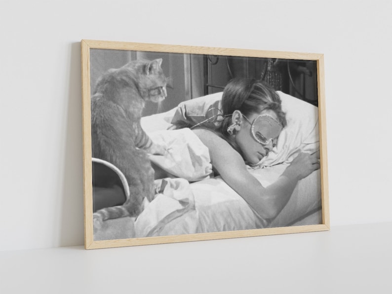 May include: A black and white photograph of a woman sleeping in bed with a cat sitting on the bed next to her. The woman is wearing a sleep mask and has her hair pulled back. The cat is looking at the camera.