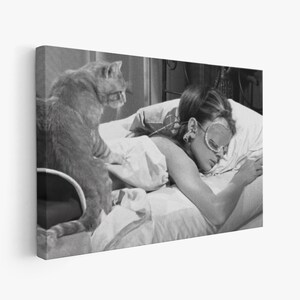 May include: A black and white photo of a woman sleeping in bed with a cat sitting on the edge of the bed. The woman is wearing a sleep mask and has her arm resting on a pillow. The cat is looking at the camera.