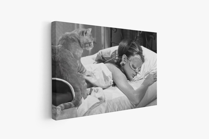 May include: A black and white photo of a woman sleeping in bed with a cat sitting on the edge of the bed. The woman is wearing a sleep mask and has her arm resting on a pillow. The cat is looking at the camera.