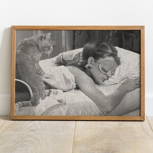 May include: Black and white photo of a woman sleeping in bed with a cat sitting on the bed behind her. The woman is wearing a sleep mask and has her hair pulled back. The cat is looking at the camera.