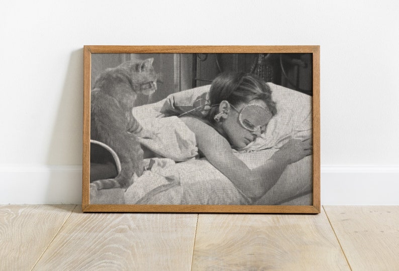 May include: Black and white photo of a woman sleeping in bed with a cat sitting on the bed behind her. The woman is wearing a sleep mask and has her hair pulled back. The cat is looking at the camera.